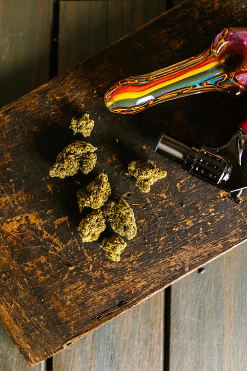 Cannabis Buds and Glass Pipe at Nature’s Wonder Dispensary Close-up of cannabis buds and a colorful glass pipe resting on a rustic wooden surface at Nature’s Wonder Dispensary.